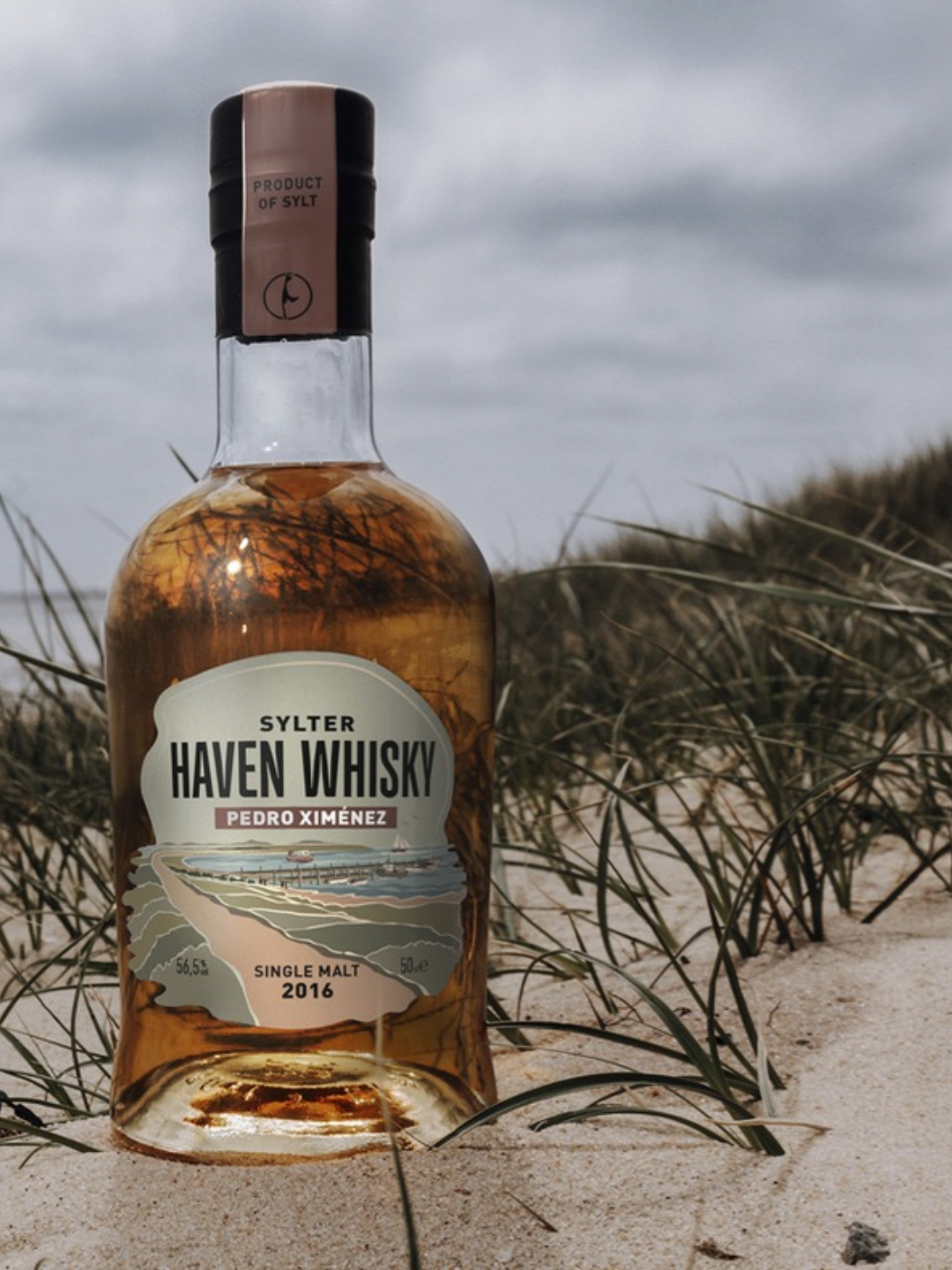 Flasche Sylter HAVEN Whisky Pedro Ximénez im hellen Dünensand zwischen Strandhafer unter bewölktem Himmel.